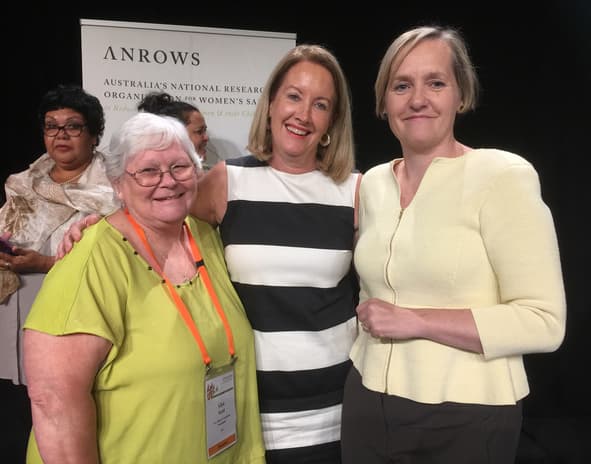 Three women smiling in front of an ANROWS banner.